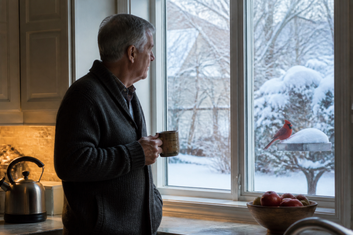 Man at a kitchen window watching a cardinal on a snowy feeder Man in his sixties with a mug at a kitchen window at dawn, a red cardinal on a snow-covered feeder just outside
