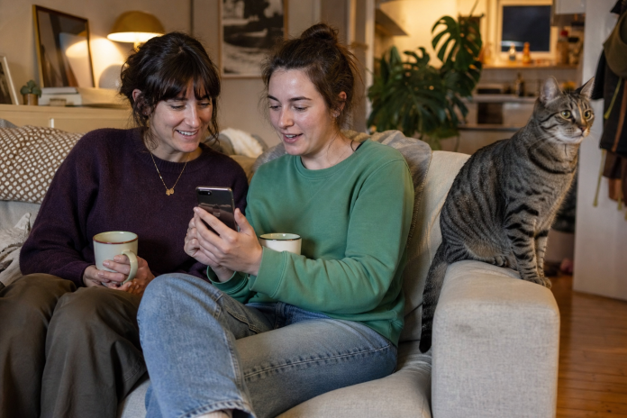 Two friends reading a text message on a sofa A woman in her late twenties showing a friend a text message on a phone, both with tea, on a sofa in evening lamp light with a cat on the armrest