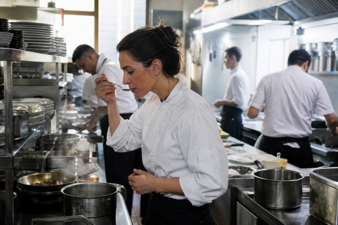 Chef tasting a sauce in a busy kitchen A woman chef pausing to taste a sauce from a spoon in a busy restaurant kitchen with steam and stainless steel