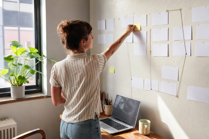 Manifesting your desires with a real plan Woman in her thirties at a small home-office desk placing a sticky note on a wall of index cards arranged as a plan, laptop open, houseplant nearby