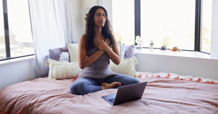 A woman sits cross-legged on a bed with her hands over her chest and a laptop nearby.