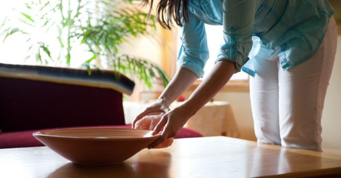 Arranging a room with intention A woman in a blue shirt arranges a shallow bowl on a wooden table beside a window.