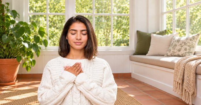 A quiet heart-centered reflection A woman in a white knitted sweater rests her hands over her chest in a bright sunroom.