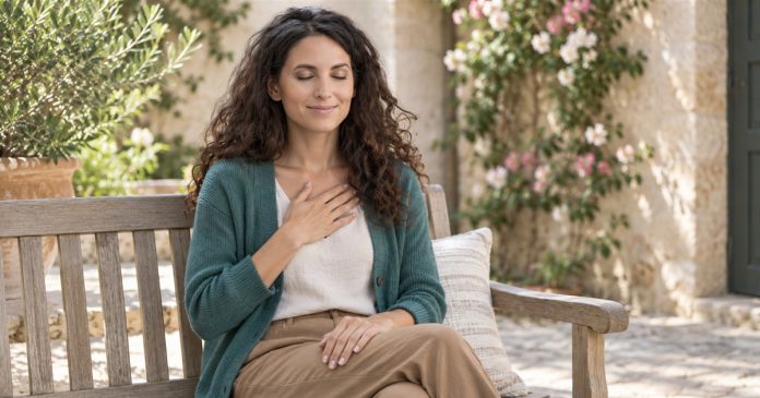 A dark-curly-haired woman sits on a garden bench with her eyes closed, one hand on her chest and the other on her lap.