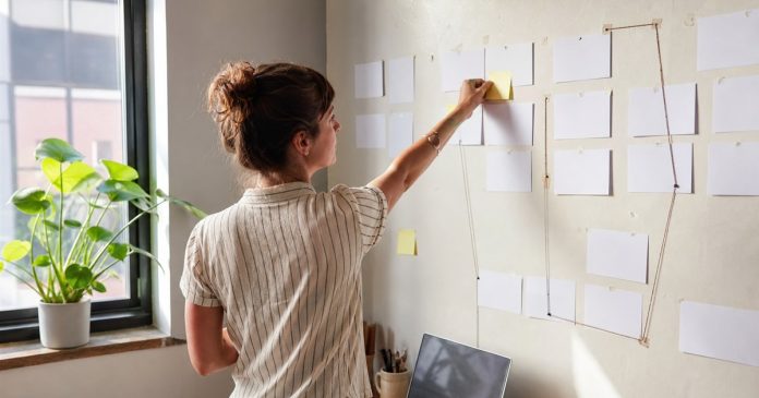 Manifesting intention board A woman arranging notes on a wall beside a wooden desk and an open laptop.